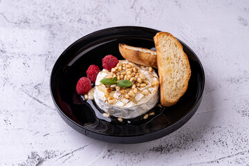 An elegant plate holds a sumptuous cheese topped with pine nuts and drizzled with honey, accompanied by fresh raspberries and crispy toasted bread, perfect for gourmet moments