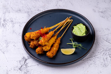 Succulent skewers of deep-fried delicacies rest on a sleek black plate, garnished with fresh herbs, lime, and accompanied by a zingy green dipping sauce, inviting a flavorful culinary adventure