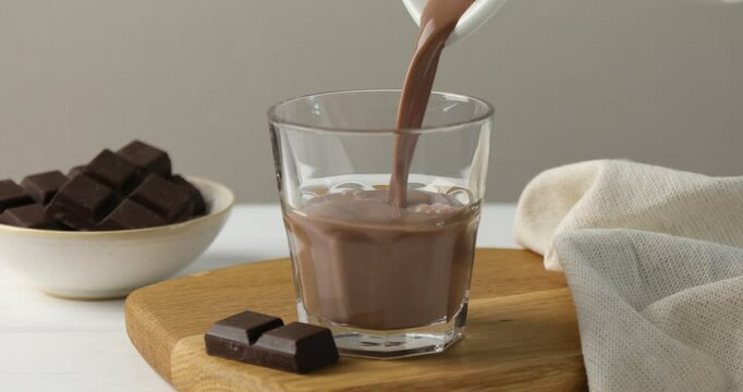 Woman pouring tasty chocolate milk into glass at white wooden table, closeup