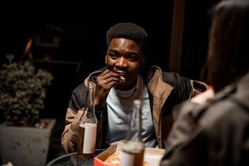 Young man enjoying snack outdoors at night with friends while having drinks and conversing