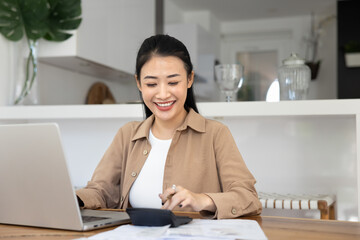 Smiling Asian woman managing her personal finances at home, paying bills online, budgeting and...