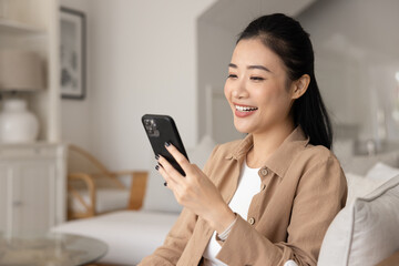 Happy Asian woman engaged in remote digital communication using modern smartphone, relaxing alone...