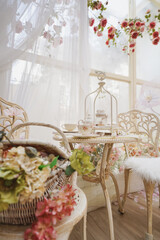 Afternoon Tea. A set of an afternoon tea by the window with cookies, biscuits in the glass jar and vintage teacups on the table.  Foreground is a blurry basket of flowers. Soft window light.