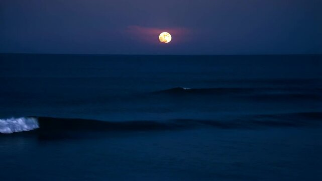 Full moon at night over the ocean with small waves under a dark purple and blue sky at the beach
