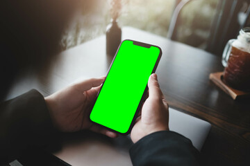 User holding smartphone with green screen at dark wooden desk with iced coffee in background,...