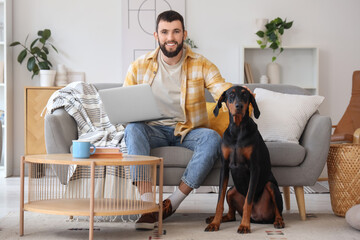 Young man with laptop and Dobermann dog sitting on sofa at home
