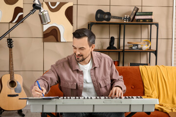 Young happy male composer writing notes while playing synthesizer in music studio