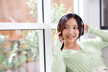 Happy young asian woman wearing headphones listening music while sitting on sofa with fun and enjoy...