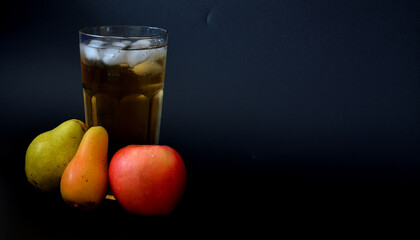 Apple and pear juice in a tall faceted glass with ice on a black background, next to ripe fruits.