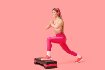 Sporty young woman with headphones exercising on aerobic stepper against pink background