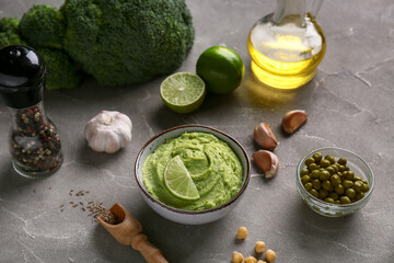 Bowl of tasty peas hummus with limes, broccoli and garlic on grey background