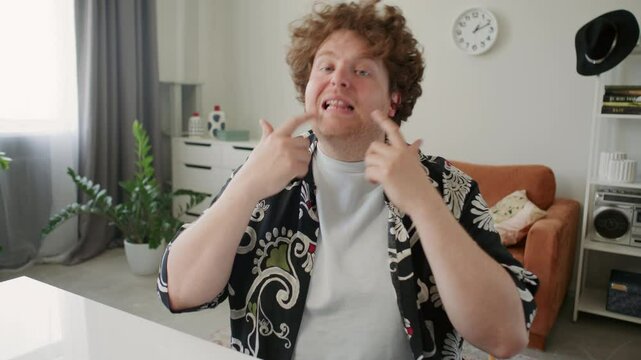 Young male teacher sitting at desk in living room, talking to camera and demonstrating breathing technique during online speech coaching session