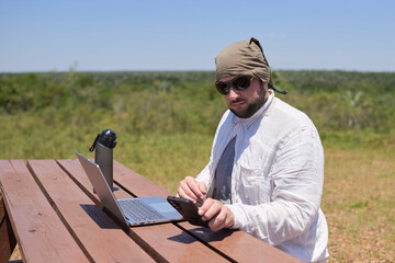 Young Hispanic man checks his phone while working on his laptop at an outdoor table on a sunny day....