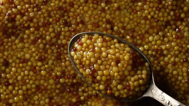 French dijon mustard in a metal spoon, top view on grains mustard background, rotates. Gastronomy concept, organic food. Close up