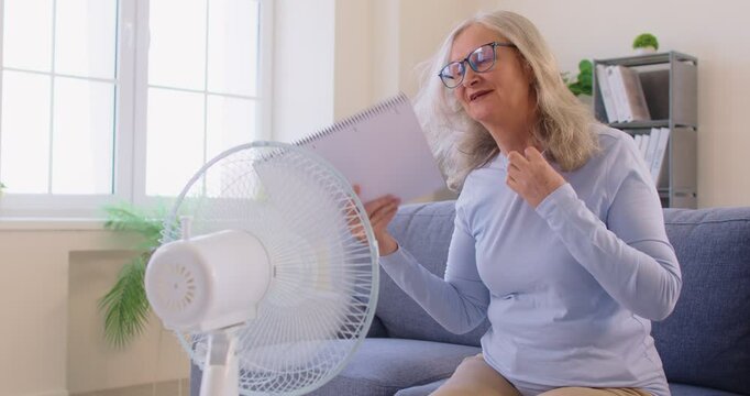 Displeased elderly woman feeling hot, suffering from high temperature during summer, using electric fan to cool herself, cannot stand such heat, needs air conditioning. Oh, its boiling here