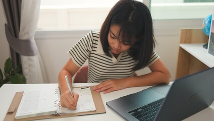 Asian girl studying at home with focus, writing in a notebook while using a laptop, representing homeschooling, distance learning, and productive education in a calm space.