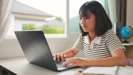 Asian girl using laptop for online learning at home, representing distance education, homeschool, and digital skills development in a calm and focused study environment.