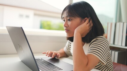 Asian teenage girl attentively watching an online class on a laptop at home, representing focused digital learning, homeschool, and remote education in a calm study space.