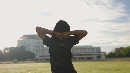 Asian teenage girl stretching arms outdoors in the morning sunlight at a sports field, representing...