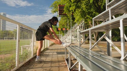 Asian teenage girl stretching her leg before workout on an outdoor sports field, promoting healthy lifestyle, fitness, and self-care through physical activity in natural sunlight.