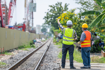 Two construction workers wea safety vests and helmets standing near railway tracks discussing work plan outdoors du daytime with lush greenery and nearby buildings in the background