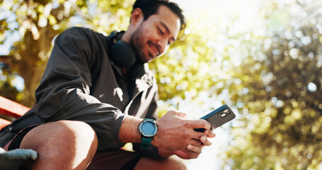 Phone, fitness and man on park bench for break, training stats and text message online in low angle. Mobile, smile and athlete resting outdoor with app for workout schedule or track health progress