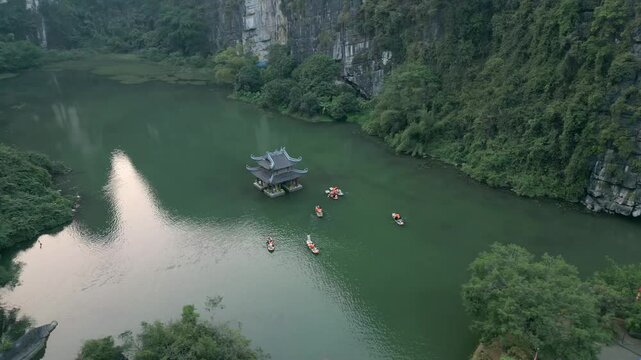 Aerial view of floating pagoda on green river, boats circling temple, lush karst mountains and cliffs form stunning natural landscape in Ninh Binh, Vietnam.
