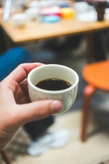 Hand holding a cup of coffee with a blurred background of a table and chairs