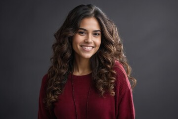 Charming young woman with long hair and a warm smile, dressed in maroon, standing before a gray background.

