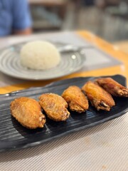 Golden-brown crispy fried chicken wings served on a sleek black plate, a popular appetizer or snack, with a blurred background of white rice, hinting at a full meal