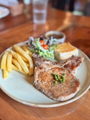 Juicy grilled pork chop steak served with golden french fries, a fresh garden salad, and toasted garlic bread on a rustic wooden table, a classic Western comfort meal.