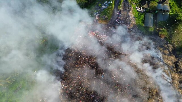 Aerial drone footage of open trash burning at a rural landfill. Smoke rises into the air. Top down view