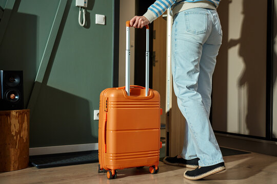 Person leaving home with orange suitcase. Female traveler start trip with luggage. Woman in casual outfit stands near door holding suitcase handle. Travel concept