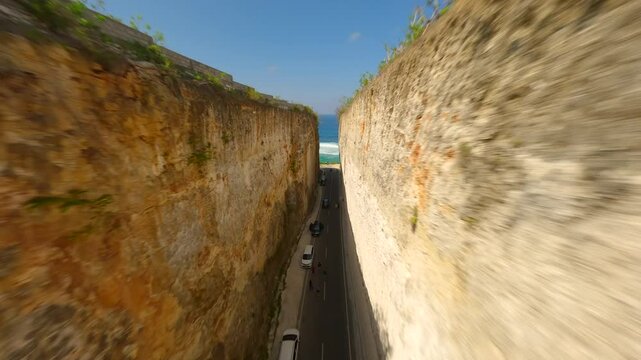 Immersive FPV drone flight along coastal road carved between limestone cliffs and turquoise waters at Batu Barak Beach, Bali, Indonesia.