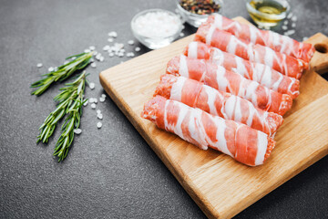 Raw meat rolls wrapped in bacon on wooden board with spices