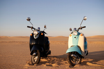Two retro scooters—blue and black—stand still in the golden Namib Desert. A quiet moment of...