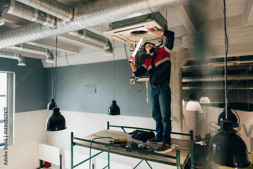 Poster worker repairs air conditioning unit while standing on scaffold in modern workspace during daylight 
