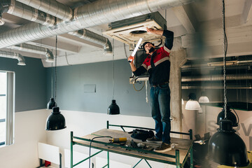 Worker repairs air conditioning unit while standing on scaffold in modern workspace during daylight hours