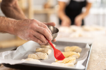Close up of baker hands brushing uncooked dough pieces on baking sheet with oil before baking in oven