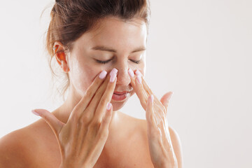 Close-up of young woman gently cleansing her face with fingertips, promoting facial skincare routine, softness, and self-care with natural light on white background.