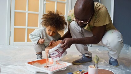 African American man engaging in painting activity with his young child in living room, both...