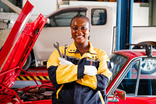 Smiling female auto mechanic in yellow and black uniform holds wrenches confidently in a repair garage, showcasing car maintenance skills and empowerment in the automotive service industry.