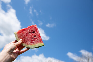 A slice of watermelon in a hand against a blue sky with white clouds. A Piece of summer, a bright and juicy summer background 