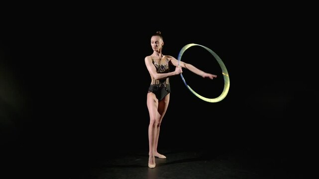 Professional skillful attractive female athlete in elegant leotard performing rhythmic gymnastics elements, passing whole body through hoop on black background.