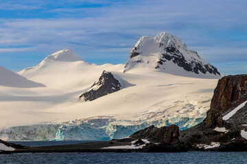 Snow covered mountain peak, early morning, Antarctic Peninsula. Rocks exposed on snowy slope. Blue ice visible. Blue sky, clouds in background. 
