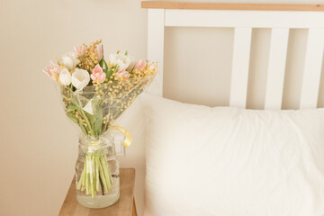 Bouquet of fresh white and pink tulips with yellow mimosa in a vase on a wooden nightstand in a bedroom. Spring flower arrangement for home interior with light colors next to a bed.