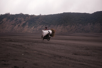 a person on a motorcycle through a volcanic environment, with a bundle of grass on the back. Java, Indonesia - 19th March 2025
