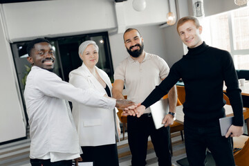 Diverse team of professionals joining hands in modern office workspace highlights collaboration,...