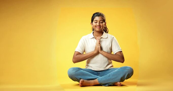 Girl Doing Mudra or Pranam, Indian Asian Schoolgirl Sitting Cross-Legged with Eyes Closed on Yellow Seamless Background Practicing Meditation with Hand Gesture for Calm Mindfulness and Inner Peace