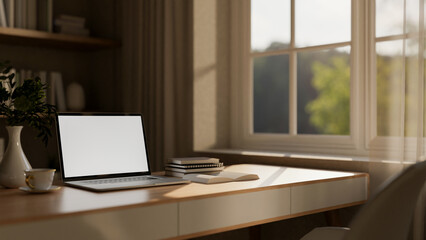 White screen laptop with books and coffee cup on wooden table next to the window with sheer curtain.